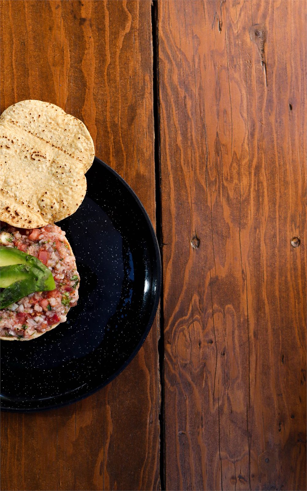 Tostada de ceviche sobre plato negro en mesa de madera, vista cenital